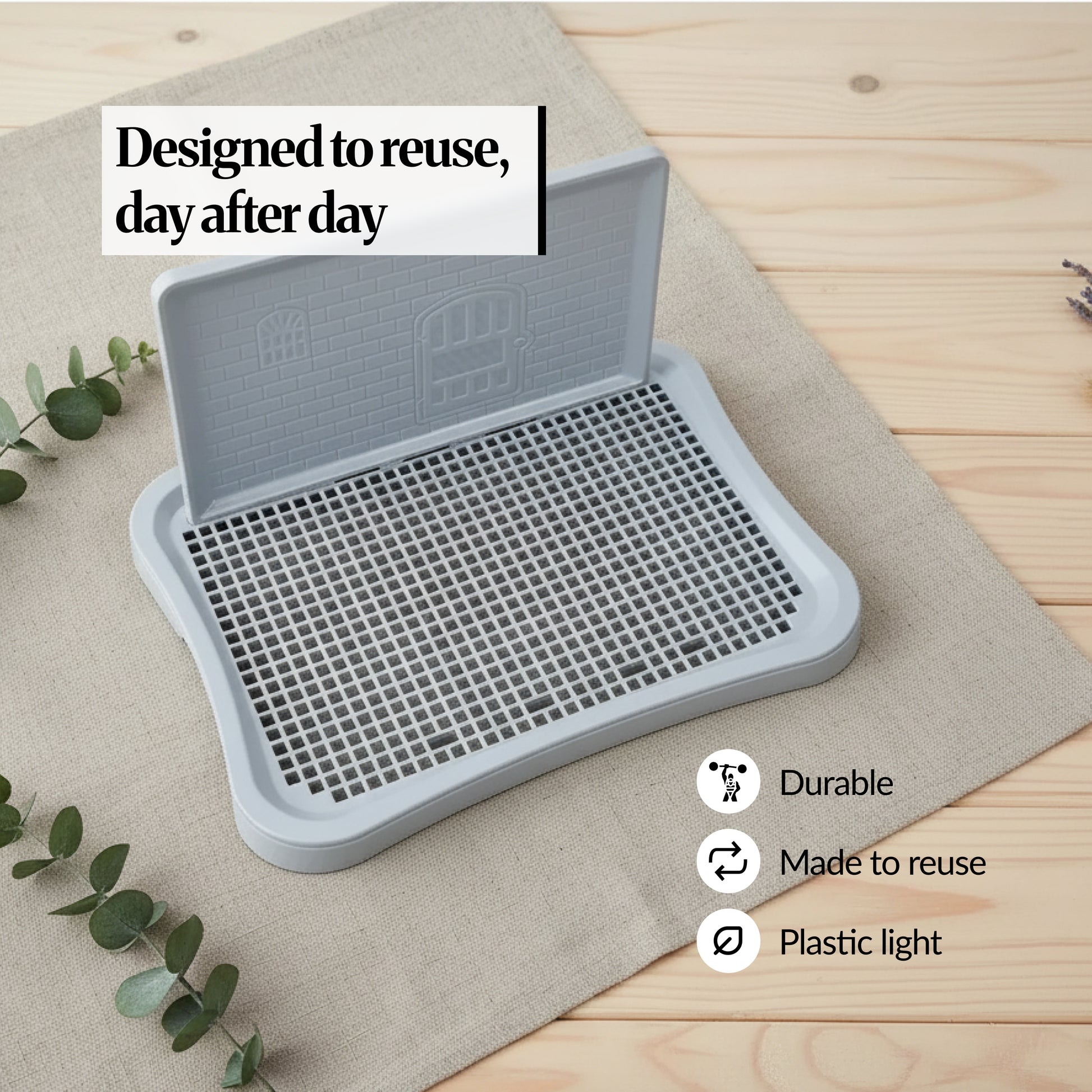 Reusable indoor dog potty tray with upright splash-guard wall and raised grid, shown on linen cloth with “Designed to reuse, day after day” text and durability icons.