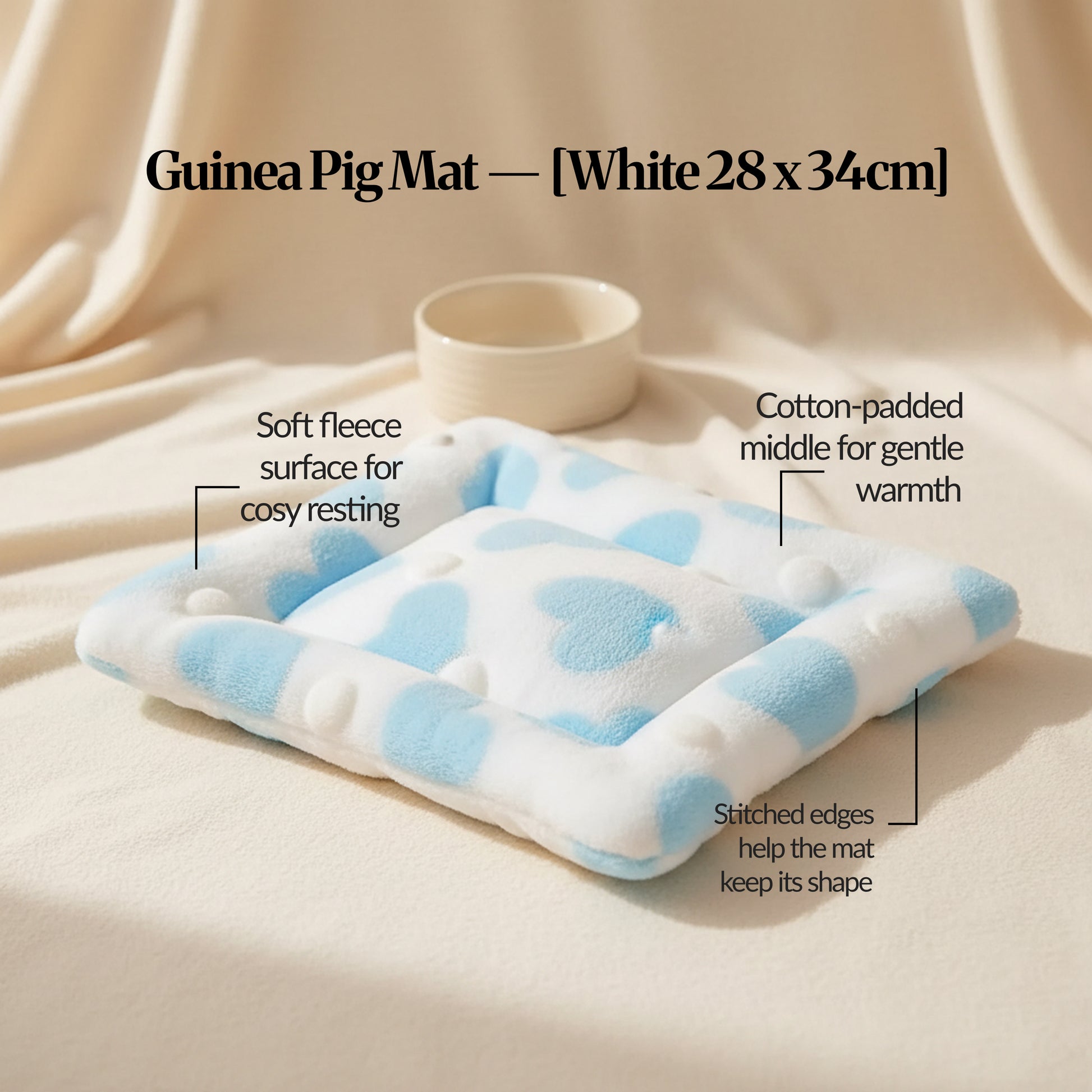 Guinea pig bed mat (White, 28×34cm) on a cream fabric backdrop with a small bowl, highlighting soft fleece, padded centre and stitched edges.