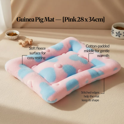 Pink guinea pig bed mat (28 × 34 cm) with soft fleece surface and stitched edges, shown on a light floor with small pet bowls in the background.