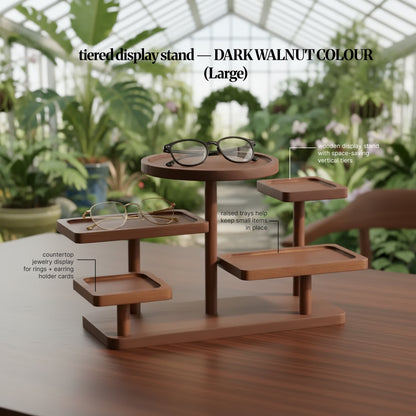 Dark walnut colour “Large” tiered display stand in a greenhouse setting, staged with eyeglasses and feature callouts for space-saving vertical tiers and raised trays that keep small items in place.