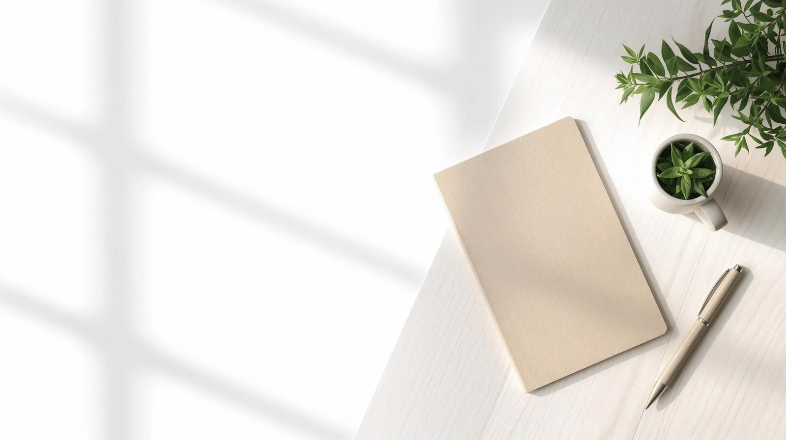 Soft, minimalist overhead scene showing a closed beige notebook, pen and a small green plant in a mug on a pale wooden desk, with gentle window light casting shadows across a clean white background.
